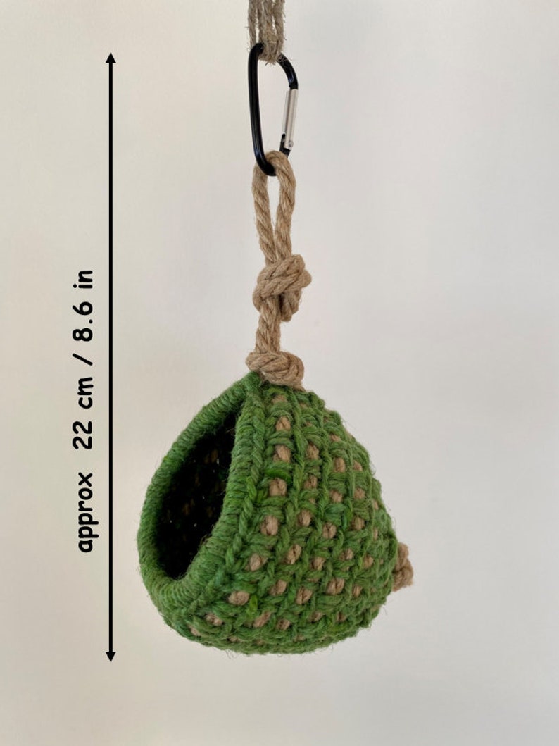 May include: A green and tan crocheted hanging pod with a circular opening, suspended by a thick tan rope and a black metal carabiner. The item measures approximately 8.6 inches in height, as indicated by the text and arrow on the left side.