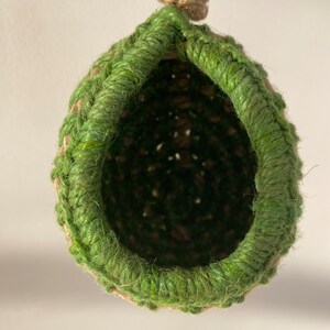 May include: A hanging, teardrop-shaped nest crafted from natural tan jute twine, featuring a thick, vibrant green fiber rim around the circular opening. The structure is suspended by a sturdy, knotted jute rope loop at the top, showing a deep, dark interior.
