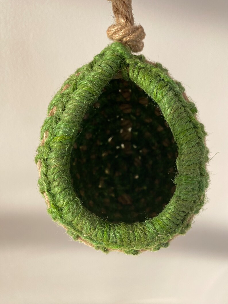 May include: A hanging, teardrop-shaped nest crafted from natural tan jute twine, featuring a thick, vibrant green fiber rim around the circular opening. The structure is suspended by a sturdy, knotted jute rope loop at the top, showing a deep, dark interior.