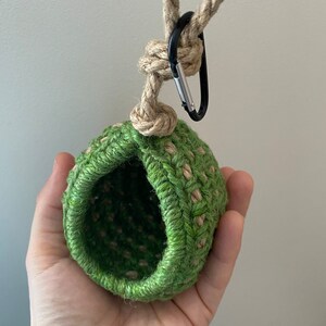 May include: A small, round, crocheted pouch made from green and tan jute twine. The pouch features a wide, open circular top and is attached to a thick, knotted jute rope loop. A black metal carabiner is clipped onto the rope for hanging.