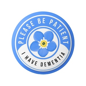 Può includere: Un bottone rotondo azzurro con la scritta bianca "PLEASE BE PATIENT I HAVE DEMENTIA". Al centro, un fiore blu non ti scordar di me con centro giallo.