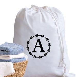 Può includere: Un sacco per la biancheria bianco con chiusura a coulisse. La borsa presenta un design a ghirlanda nera con la lettera "A" al centro. Un cestino di vimini con asciugamani blu e bianchi piegati è in primo piano.