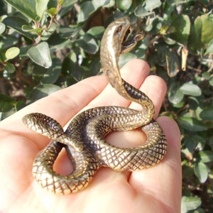 May include: A bronze-colored snake figurine with an open mouth and raised head. The snake is coiled, with detailed scales visible. The sculpture is held in a hand against a backdrop of green foliage.