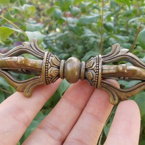 Könnte beinhalten: Ein bronzefarbener Vajra, ein Ritualobjekt mit aufwendigen Details. Die zentrale Kugel wird von dekorativen Elementen und spitzen Enden flankiert. Das Objekt wird vor einem Hintergrund aus grünem Laub gehalten.