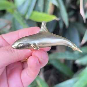 May include: A small, gold-colored dolphin figurine. The dolphin has a sleek, streamlined body with a dorsal fin and flippers. The figurine is held in a hand against a blurred green background.
