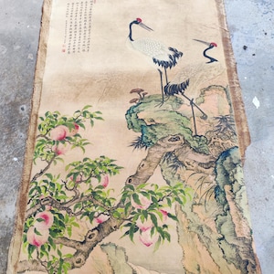 Puede incluir: Una antigua pintura china sobre papel envejecido. Dos grullas coronirrojas se alzan sobre una formación rocosa, con un melocotonero en primer plano. La pintura presenta pinceladas detalladas y caligrafía.