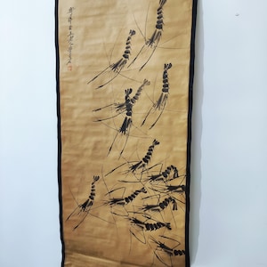 Puede incluir: Una pintura en rollo vertical que representa un grupo de camarones en tinta negra sobre un fondo marrón. La obra de arte tiene una estética asiática tradicional, con caligrafía en la esquina superior izquierda y un borde negro.