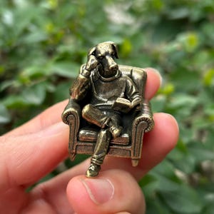 Könnte beinhalten: Eine bronzefarbene Figur eines hundeköpfigen Wesens, das in einem kleinen Sessel sitzt und ein Buch liest. Die Figur trägt einen Pullover und eine Hose. Der Sessel hat detaillierte Armlehnen und ein gemustertes Design.