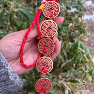 Könnte beinhalten: Ein dekoratives Hängeornament mit fünf roten und goldenen chinesischen Münzen, die auf einem Holzdübel mit roter Kordel aufgefädelt sind. Die Münzen haben komplizierte chinesische Schriftzeichen und sind mit roter Schnur gebunden. Das Ornament hat eine rote geflochtene Kordel zum Aufhängen.
