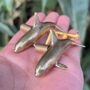 May include: A pair of gold-colored dolphin cufflinks. The cufflinks are detailed with fins, tails, and small eye details. The dolphins are connected by a gold-colored bar.