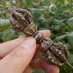 May include: A bronze-coloured vajra, a ritual object, is held against a backdrop of green foliage. The vajra features intricate, ornate detailing with a central bulbous section and flared ends. It is a symbol of enlightenment.