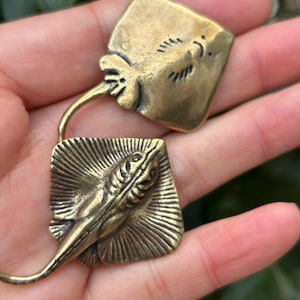 Puede incluir: Dos colgantes de latón con forma de manta raya. Cada colgante presenta un diseño detallado de manta raya con cuerpo cuadrado y cola curvada. Los colgantes están conectados por un lazo de latón curvado. El latón tiene un acabado envejecido y antiguo.