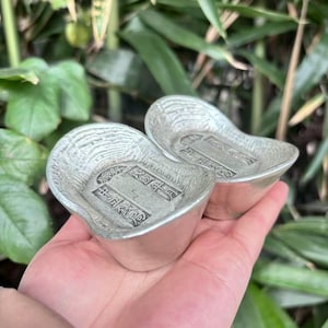 Puede incluir: Se muestran dos lingotes de plata, cada uno con forma de barco. Los lingotes tienen grabados detallados y se sostienen en una mano sobre un fondo de follaje verde. Los lingotes probablemente son de plata.