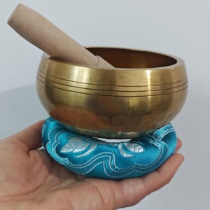 May include: A golden singing bowl with a wooden striker rests on a teal cushion with a silver floral design. The bowl has horizontal etched lines and is held in a hand.