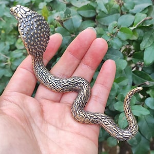 May include: A gold-coloured metal snake figurine with a textured surface. The snake is coiled with its head raised and has a detailed head with scales.