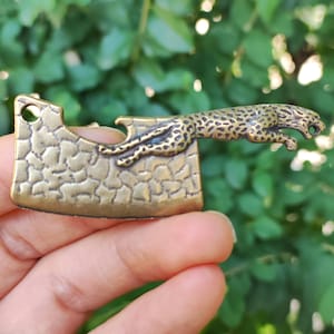 May include: A bronze-colored metal cleaver-shaped pendant with a leaping jaguar design. The cleaver has a textured surface and a hole for attaching to a chain or cord. The jaguar is detailed with spots and a snarling expression.
