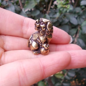 Puede incluir: Pequeña y detallada figura de latón de un perro-león mítico sentado, un Foo Dog. La escultura es de color marrón dorado con intrincadas tallas, incluyendo una melena fluida y una bola bajo su pata. La figura se sostiene en una mano.