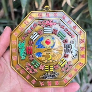 May include: An octagonal, gold-colored metal pendant with intricate designs and Chinese characters. The center features a yin-yang symbol with a sun, moon, and cloud motif. Decorative elements include a dragon, phoenix, and other symbolic figures.