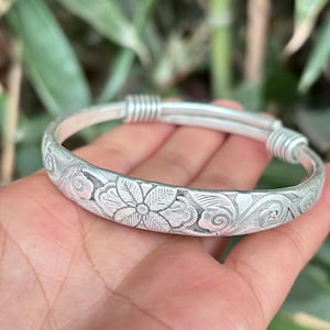 以下が含まれることがあります： 銀色のバングルブレスレット。彫刻された花の模様が特徴です。ブレスレットは、花と渦巻く葉の細かい模様が特徴です。ブレスレットは調節可能で、滑らかで磨かれた仕上がりです。デザインはブレスレットの中央に配置されています。