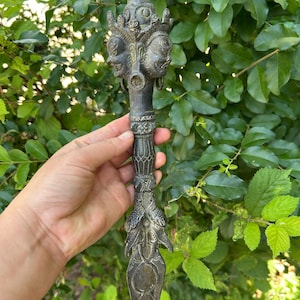 May include: A dark metallic ritual dagger featuring a pointed blade, a handle with intricate geometric carvings, and a pommel adorned with three sculpted faces topped by a small animal figure. The object has a weathered, aged patina finish.