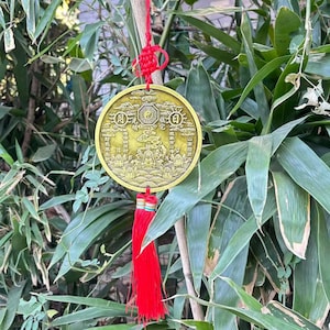 Könnte beinhalten: Ein runder, goldfarbener Metallanhänger mit chinesischen Schriftzeichen und einem zentralen Yin-Yang-Symbol. Er hängt an einer roten Kordel und Quaste vor grünem Bambuslaub. Der Anhänger hat ein aufwendiges Design.