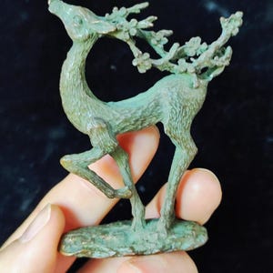 May include: A small, verdigris-coloured deer figurine with floral antlers. The deer is posed with its head raised and mouth open. The figurine is held in a person's hand, and the base is oval-shaped.