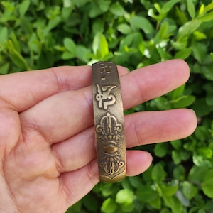 May include: A bronze-colored metal bracelet with embossed religious symbols. The bracelet features intricate designs, including what appears to be script and a crown-like emblem. The background is a blurred view of green foliage.