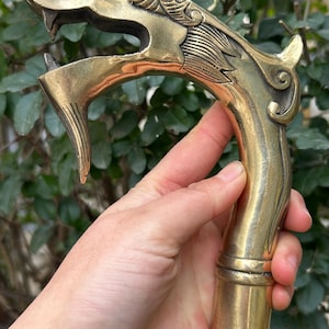 Puede incluir: Bastón de latón con diseño de cabeza de dragón. La cabeza del dragón está en la parte superior del bastón, con detalles intrincados en la cara y la melena. El bastón tiene un mango curvo y un acabado liso y pulido.