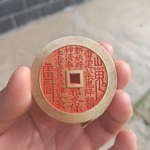 May include: A circular, gold-toned coin featuring red Chinese characters and a square aperture at its centre. The coin is held in a hand, with a blurred backdrop of a grey wall and a light-coloured surface.