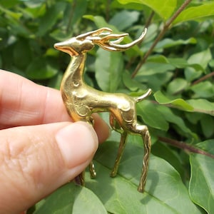 May include: A gold-colored deer figurine with detailed antlers and slender legs. The deer is held against a backdrop of green leaves, highlighting its metallic finish. This decorative item could be a brooch or a small sculpture.