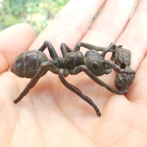 May include: A bronze-colored ant figurine, intricately detailed with six legs, antennae, and a segmented body. The ant is depicted carrying a small object, possibly food. The sculpture is held in a hand, showcasing its size and craftsmanship.