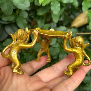 May include: A gold-colored decorative dish held by three monkey figurines. The monkeys are in a squatting position, supporting a shallow, oval-shaped dish. The background features green foliage.
