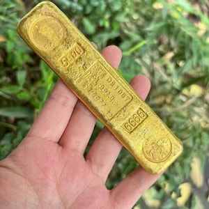 May include: A gold bar with stamped markings and a circular emblem at each end. The bar has the numbers "5000" and "6666" stamped on it, along with other text. The bar is held in a hand.