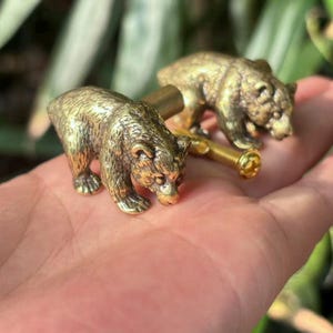 May include: Two brass bear-shaped cabinet knobs. The knobs have a textured finish, showing the bears in a walking pose. The hardware includes a brass screw for installation. The design is rustic and decorative, suitable for furniture.