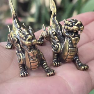 Puede incluir: Dos pequeñas y detalladas figuras de dragón de latón. Los dragones están en posición agachada con la cola levantada. El latón tiene un acabado oscuro y envejecido, que resalta las intrincadas escamas y rasgos de las criaturas míticas.