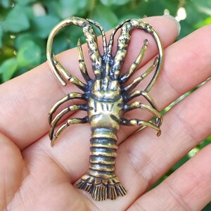Puede incluir: Figurilla de langosta de bronce detallada con una parte superior en forma de corazón. El crustáceo tiene múltiples patas y antenas, y un cuerpo texturizado. La langosta es de color oro antiguo cálido.