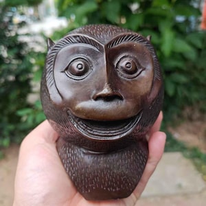 May include: A hand-carved, brown wooden sculpture of a monkey's head. The monkey has a wide, smiling mouth, large eyes, and small ears. The sculpture is held in a person's hand against a blurred green background.