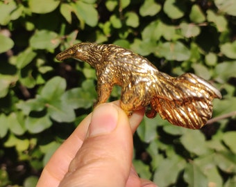 May include: A gold-coloured anteater figurine with detailed fur texture. The anteater is in a standing position, with its tail curved upwards. The background is a blurred view of green foliage.