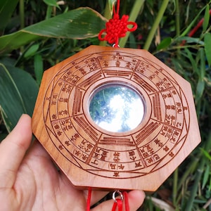 Könnte beinhalten: Achteckiger Holzspiegel mit gravierten chinesischen Schriftzeichen und einer zentralen reflektierenden Oberfläche. Aufgehängt an einer roten Schnur und einem Knoten, mit natürlicher Holzmaserung. Der Hintergrund zeigt grünes Laub.