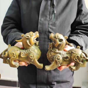 May include: Two bronze-colored, mythical creature statues. Each statue is in a crouching position with an open mouth, horns, and intricate detailing. The statues are held in front of a dark jacket.