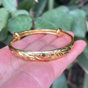 May include: A gold-colored bangle bracelet with an engraved floral pattern. The adjustable bracelet is held in a hand, showcasing the intricate details of the design. The bracelet's surface reflects light, highlighting its polished finish.