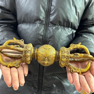 May include: An ornate golden vajra, a ritual object with intricate carvings. The central sphere is decorated with geometric patterns, and the ends feature dragon-like figures. The vajra is held in open hands against a dark background.