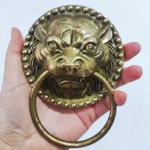 Może przedstawiać: Stary, mosiężny kołatka do drzwi przedstawiająca głowę lwa z uchwytem w kształcie pierścienia. Twarz lwa jest szczegółowa. Okrągła kołatka ma obramowanie z koralików. Mosiądz ma postarzany, antyczny wygląd.