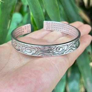 May include: A silver-colored cuff bracelet with an engraved cloud pattern. The bracelet has etched characters on the inner and outer surfaces. The bracelet is approximately 2.5 inches in diameter.