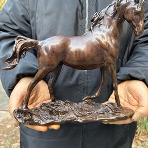 May include: A bronze sculpture of a horse in a running pose. The horse has a flowing mane and tail, and is mounted on a textured base. The sculpture is a rich brown colour and appears to be made of metal.
