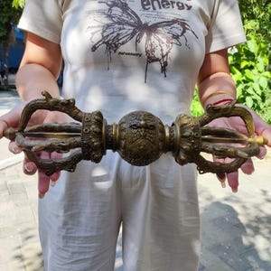 Pode incluir: Um vajra de cor bronze, um objeto ritual com detalhes ornamentados. A esfera central apresenta um design detalhado de rosto, ladeado por figuras semelhantes a dragões. As extremidades têm três pontas e o design geral é simétrico.