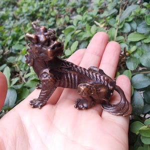 May include: A bronze-colored figurine of a mythical creature, possibly a dragon or lion, with intricate details. The sculpture is posed in a seated position, showcasing its textured body and stylized features. The figurine is approximately 4 inches long.