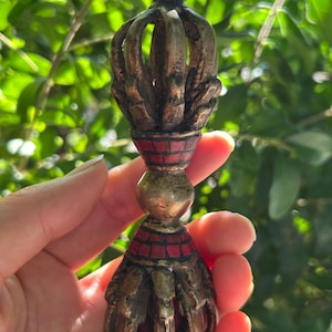 May include: A detailed, antique-style metal vajra, a ritual object. The vajra features a crown-like top, a central sphere, and a bottom with a similar design. Red and black accents are visible. The object is held against a backdrop of green foliage.