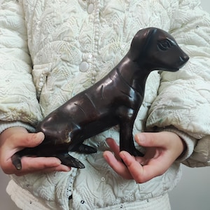 Puede incluir: Una figura de perro de color marrón oscuro, bronce. La escultura representa un perro sentado con un acabado liso y pulido. Los rasgos del perro son detallados, con orejas, ojos y una forma corporal definida. La figura está sostenida en las manos de una persona.