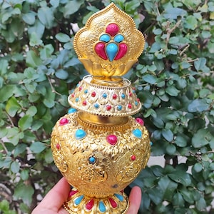 May include: A gold-colored decorative vessel with intricate embossed patterns and adorned with turquoise and red gemstones. The vessel has a lotus-shaped top and base, and is held by a hand against a backdrop of green foliage.